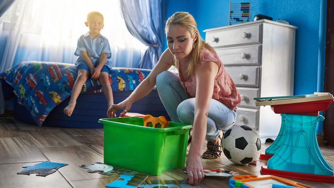 A young mother is squatting down on the floor and tidying a child’s bedroom as a young child sits on the bed watching
