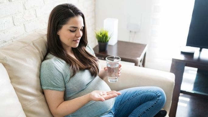 A young woman with long dark hair is smiling and holding a glass of water in one hand and probiotics tablets in the other hand