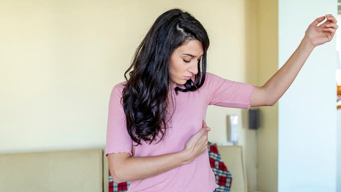 A woman with long dark hair wearing a pink top and jeans is holding one breast which looks sore and tender, which may be a sign of ovulation