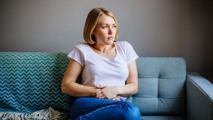 A young Caucasian woman with a concerned expression is sitting on a sofa holding her stomach