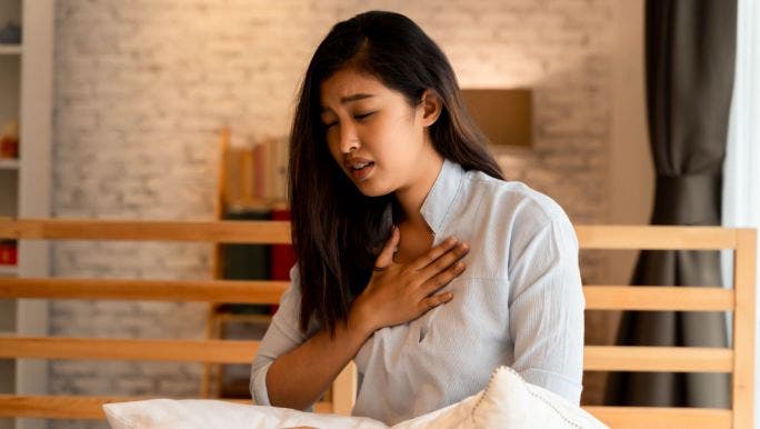 A young Asian woman is sitting in bed and holding her chest 