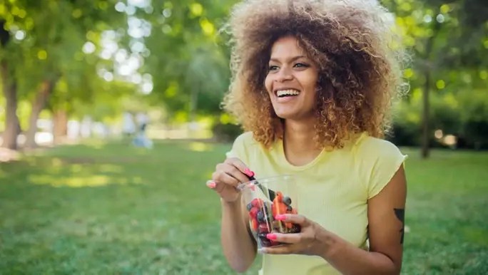 A woman in a yellow shirt sits in a park eating vegan snacks out of a container, smiling happily.