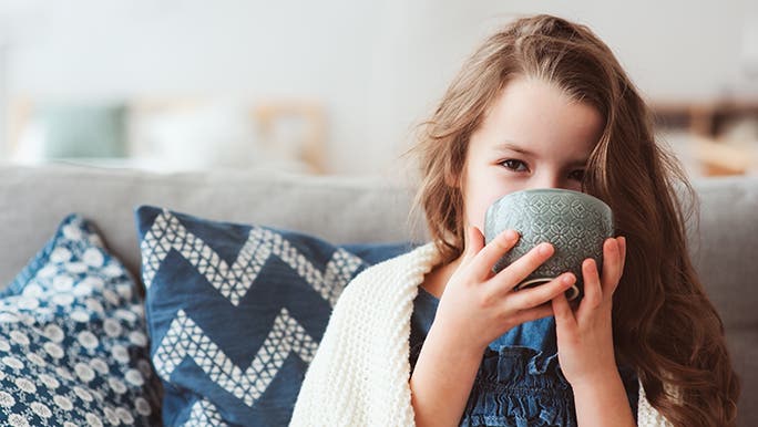 A child drinks something from a mug, she is sitting on a couch with a blanket over her shoulders. It seems as though she may be sick. 