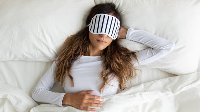 A woman with long dark hair is sleeping in a comfortable bed and wearing an eye mask