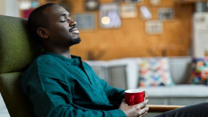 A young black man is relaxing on a sofa with his eyes closed and holding a cup of tea