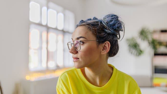 A woman in prescription glasses and a bright yellow t shirt looks away from the camera. She is in a bright white room and has colourful hair. 