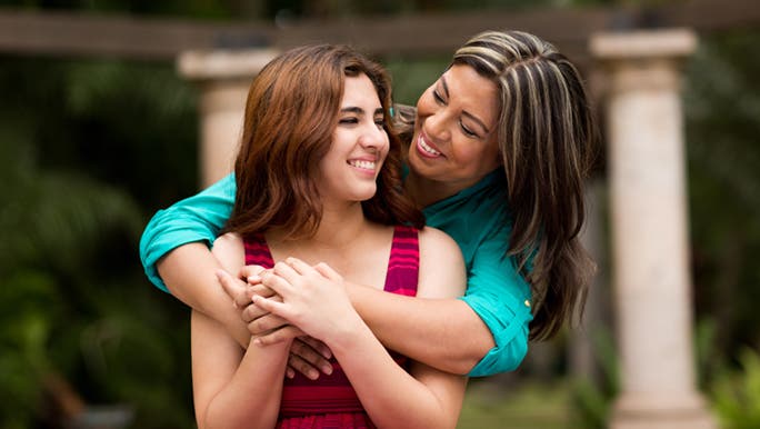 Mother and daughter are standing outside, her mother hugging her from behind as they smile. Her mother reminded her how difficult pimples can be as a teenager.  