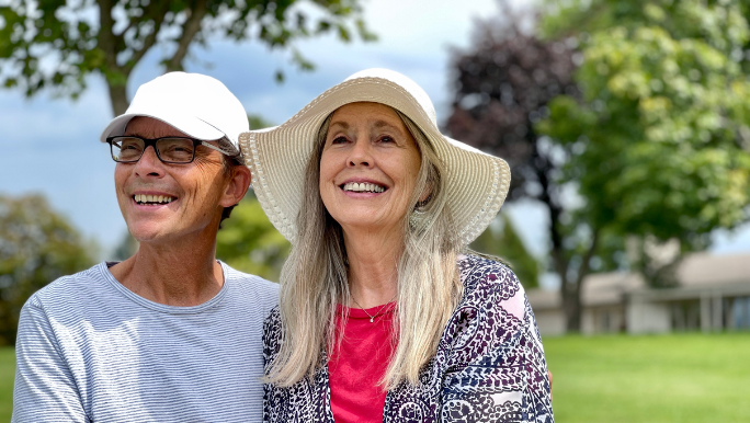 A senior couple are sitting outdoors on a sunny day smiling and wearing hats