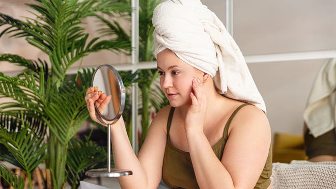 A woman wearing a towel on her head is looking into a hand held bathroom mirror