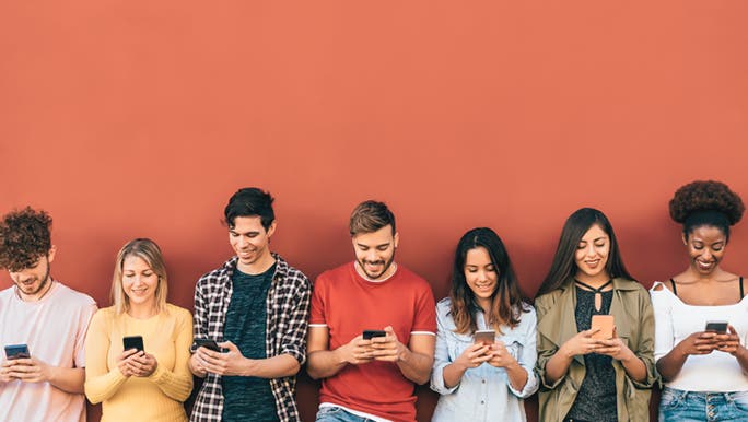 In front of a coral coloured wall, seven friends are standing in a line and using social media on their phones. 