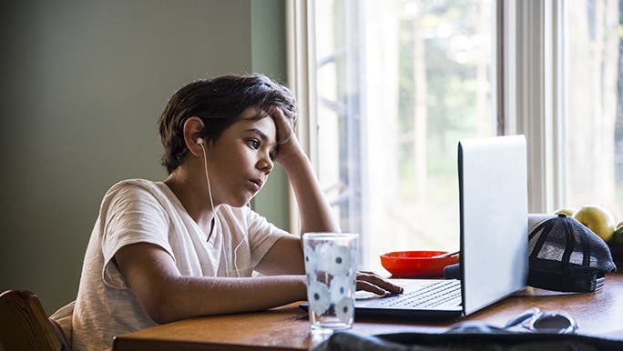 A boy sits at a desk on a laptop listening to headphones and concentrating on his homework which can be part of the recommended screen time for a child. 