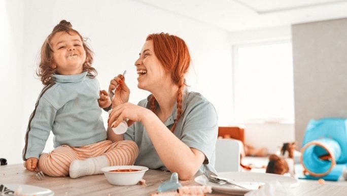 A mother is feeding yoghurt to her young daughter who is sitting on a kitchen table