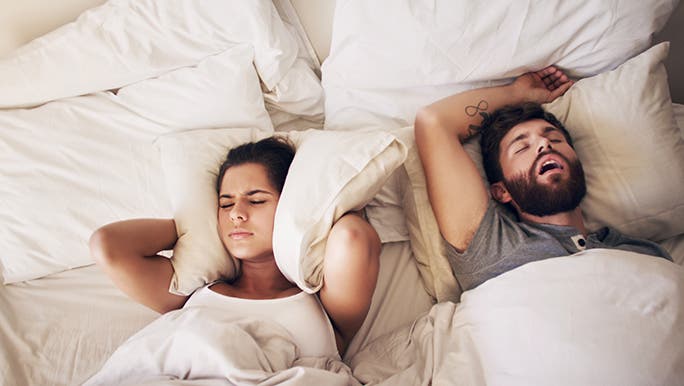 A woman and man are laying in a bed with white sheets. He is asleep with his mouth open and she is cranky-faced blocking her ears with a pillow. The woman is wondering how to stop someone from snoring. 