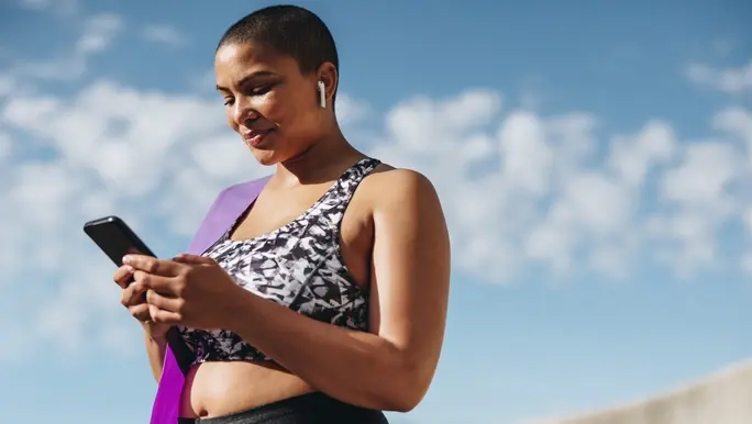 Healthy woman wearing wireless earphones taking a break from workout and checking her fitness progress on mobile phone.