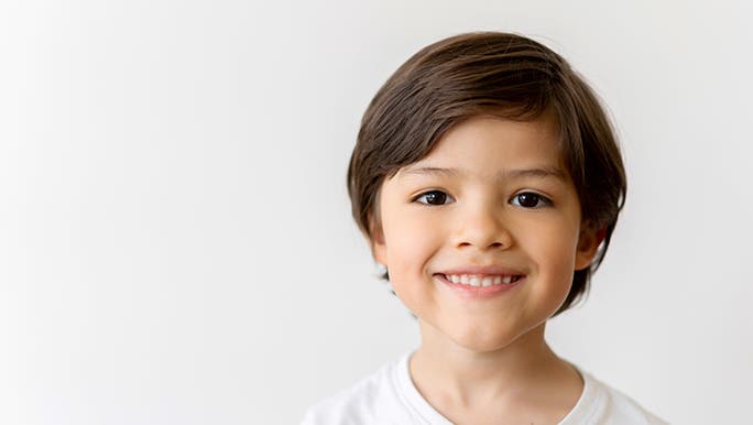 A young boy in a white shirt smiles directly into the camera. He is in front of a white wall. He looks grateful. 