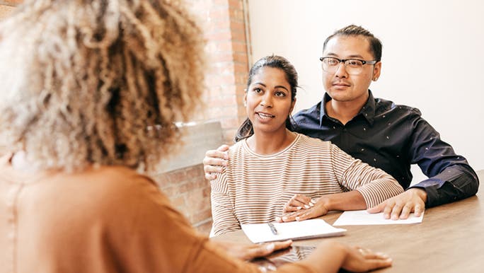An Asian man wearing a black shirt sits at a table with his arm around his female partner, who has long dark hair and dark skin, as they possibly discuss with a doctor how to optimise their fertility 