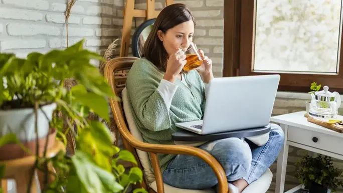 Woman sitting in an armchair sipping peppermint tea and reading about IBS and low FODMAP foods on her laptop