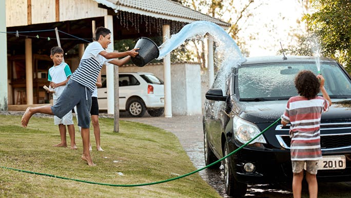 A family is getting some incidental exercise by washing the car. A teenager is throwing a bucket of water in the air, and a child is spraying the car with a hose. 