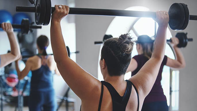 A woman in a gym class is lifting a weighted bar above her head and enjoying the benefits of lifting weights. 