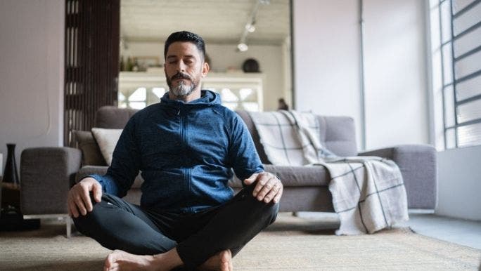 A man is sitting cross-legged on the floor practising meditation to help with stress management
