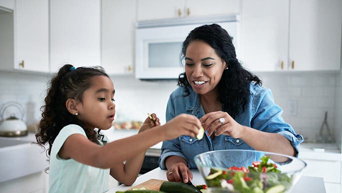 A mother prepares a salad with her daughter. The benchtop is full of seasonal fruit and vegetables. 
