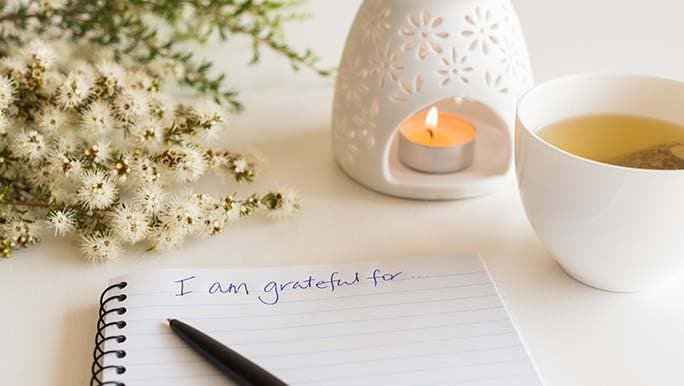 An open gratitude journal lays on a table next to an oil burner and a cup of herbal tea. The words, I am grateful for, are written in blue pen on the page.  