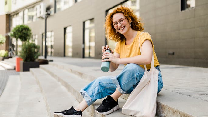 A young woman with red curly hair is sitting on stairs and holding a water bottle while smiling at the camera 