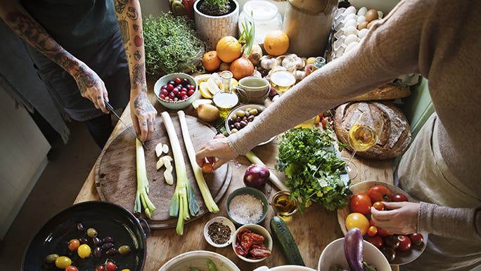 A wooden table is covered with plant based and vegan ingredients. There is someone cooking. 