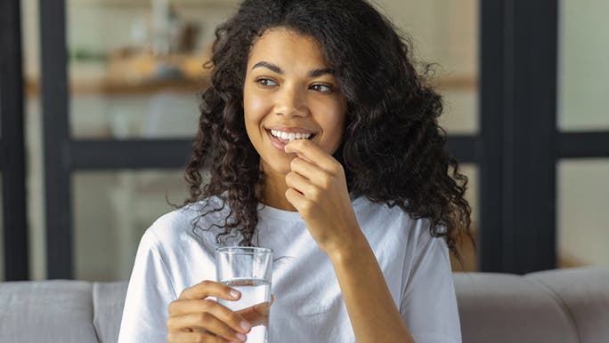 An African American woman is smiling as she holds a glass of water and takes a collagen supplement for gut health