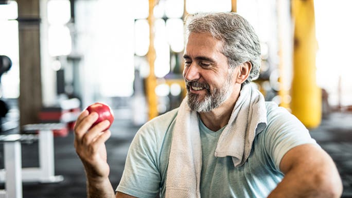 A middle aged man is in a gym smiling and holding an apple, a food considered good for gut health