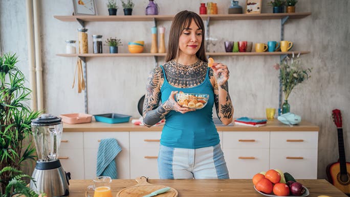 A young tattooed woman wearing a blue top with long dark hair is eating a colourful fruit salad containing bananas for gut health and other fruits