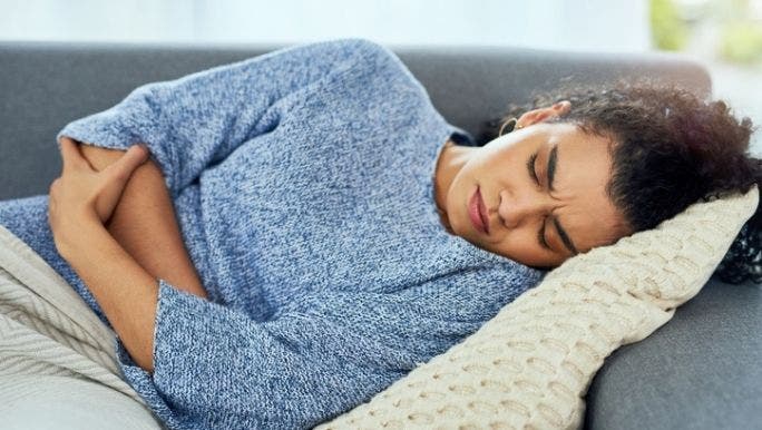 A young woman is lying on a sofa clutching her stomach and grimacing in pain