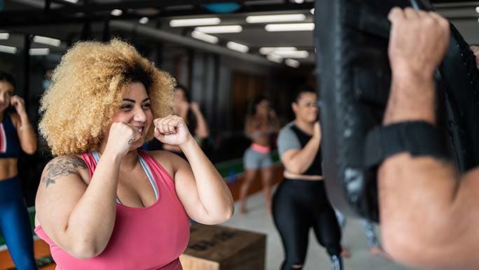 A woman smiles with her arms raised in a boxing stance about to punch a pad and knows this is how to stay motivated at the gym.