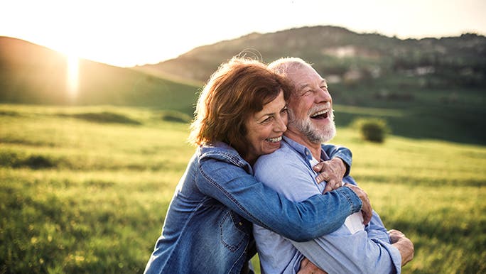 A couple embrace on a grassy hill, the sun is setting behind them and they look happy and healthy.