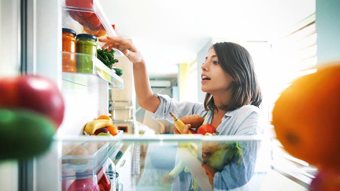 A woman has arms full of healthy food and is packing her fridge. She wants to stop eating junk food. 