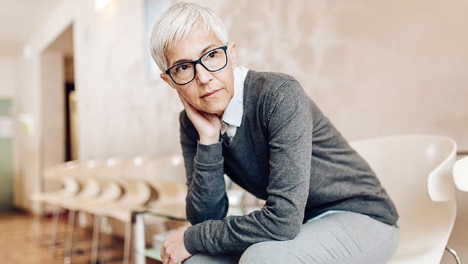 A lady with a grey pixie cut and black-framed glasses looks concerned. It seems as though she is in some sort of waiting room and may have lockdown fatigue. 