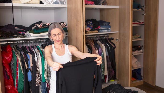 A woman dressed all in white with short hair is standing in front of an open wardrobe holding up a black top