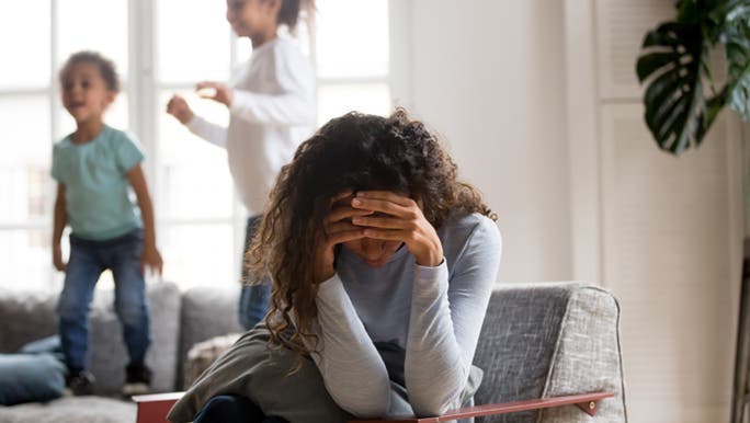 A woman sits on a chair holding her head in her hands. She is trying to deal with her stress as her children jump on the couch behind her. 