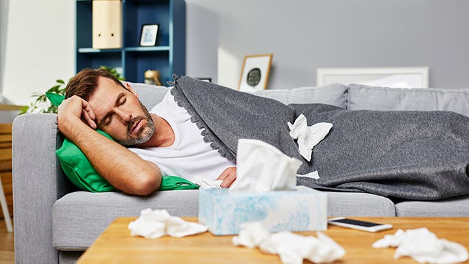 A man lies on a couch with a grey blanket and a box of tissues, pondering why he wakes up with a blocked nose.