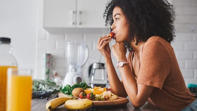 Woman leaning on the kitchen bench eating low FODMAP strawberries as she prepares a smoothie
