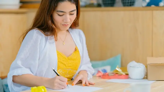 A woman is sitting at a desk writing on a card and is wondering what to write in a Galentine’s Day card.