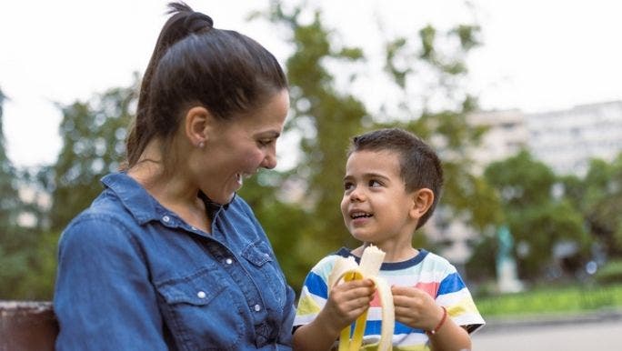 A young mother is sitting on an outdoor bench with a young boy on her lap eating a banana
