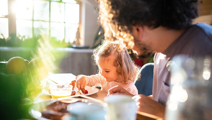 In a sun-drenched kitchen, a father watched a toddler eat her vegetables. 