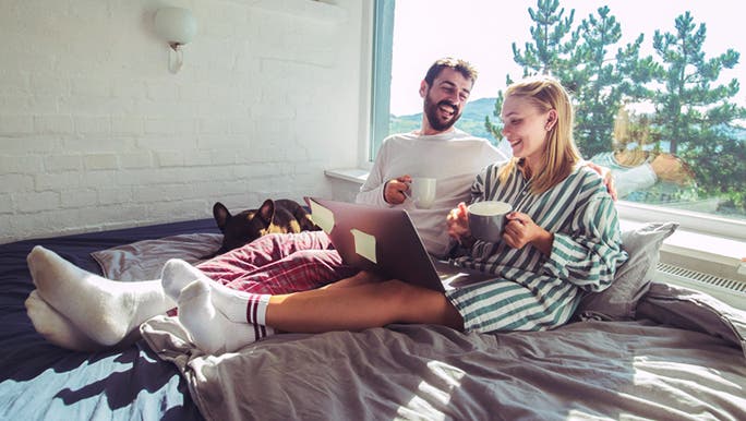 A young couple sitting up in bed smiling and holding cups of coffee