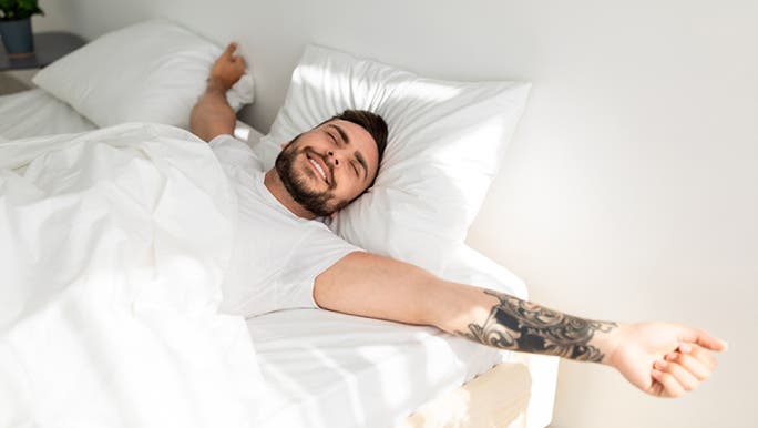 A young man with dark hair and an arm tattoo is waking up in bed