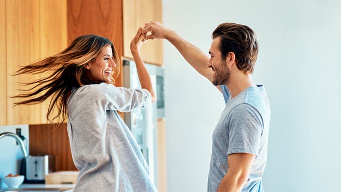 A couple are enjoying the positive mental effects of music. He is spinning her in the kitchen and they are laughing.