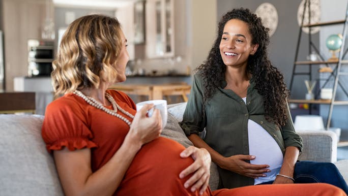 Two pregnant mothers offer emotional support to each other as they sit on a couch, with one in a red dress holding a cup of tea or coffee and both touching their bellies