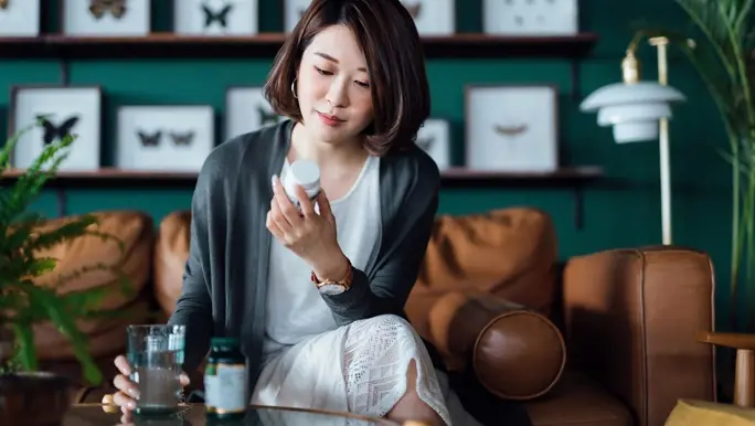 Woman taking medicines with a glass of water on the coffee table, reading the information on the label of her medication at home.