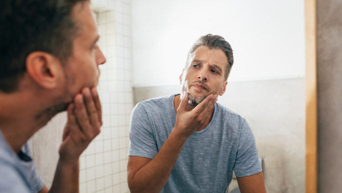 A young man with a beard is touching his face as he looks into a bathroom mirror