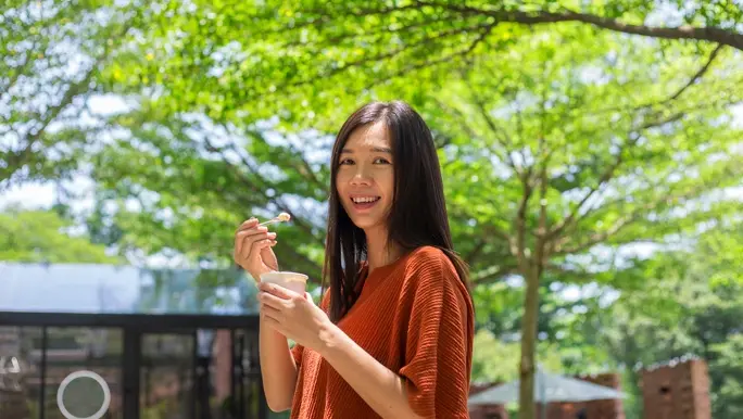 Smiling woman with long dark hair eating probiotic yoghurt in the park
